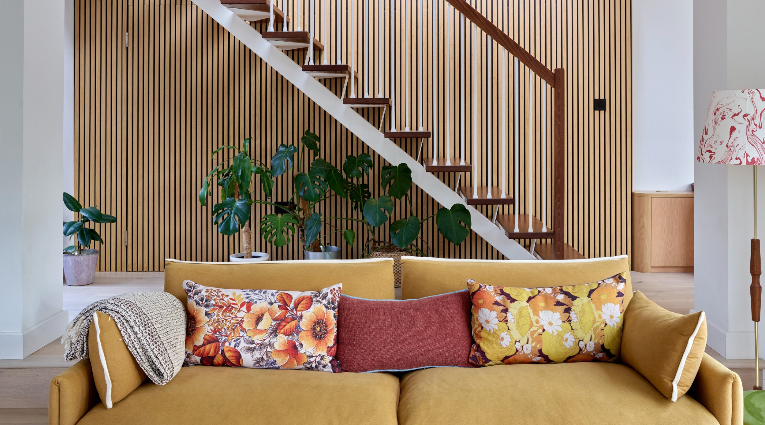 Stylish living room with yellow sofa, floral pillows, and a striking wooden wall panel feature from The Panel Hub behind a staircase.